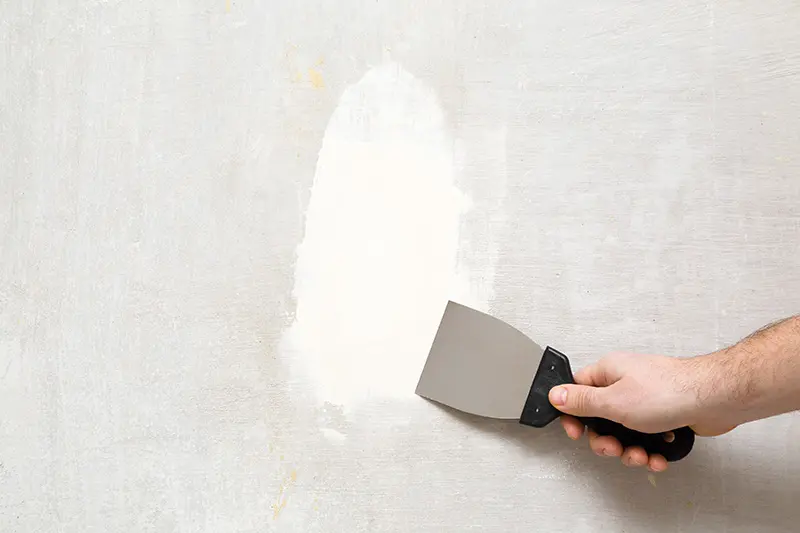 Close up of mans hand using a filling knife to smooth white filling compound over a repaired plaster wall.